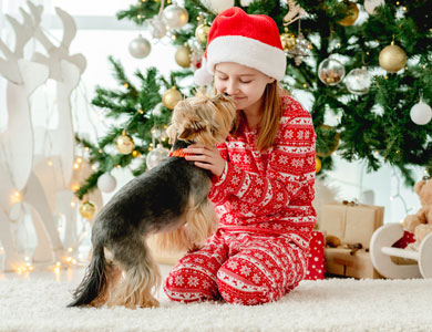 a little girl in pyjamas plays with a dog and a Christmas tree is in the background